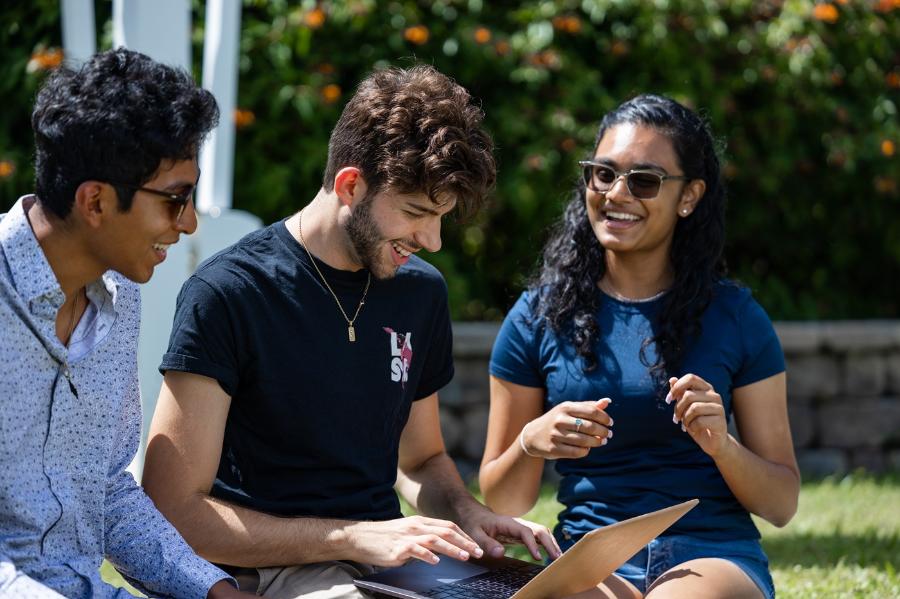 Three students sitting outside on campus; one is typing on a laptop computer.