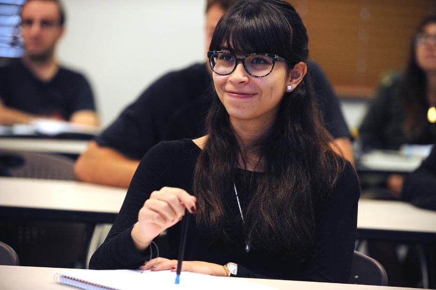 A student sits in a classroom smiling; several other students are visible in the background.