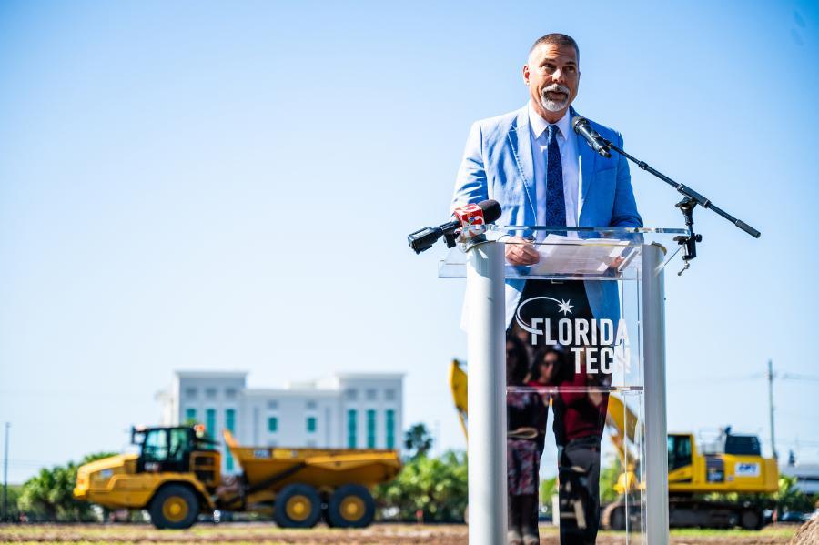 Florida Tech President John Nicklow at an acrylic podium on the site of the university's new student housing complex. Large construction machinery is behind him. Further behind him, L3Harris Commons and a blue sky.