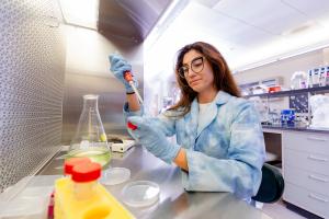 A woman student in a lab coat and protective eyewear works in the lab.