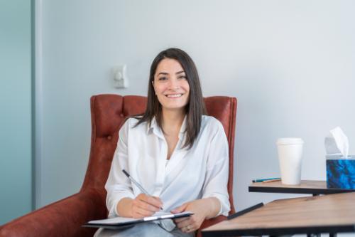 Smiling professional looking woman sitting in a chair in an office environment holding a clip board and pen.