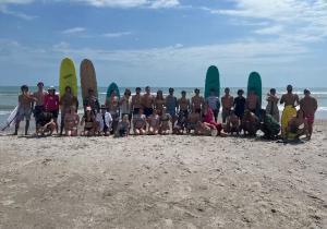 A large group of students posing at the beach with surfboards and other gear for beach activities.